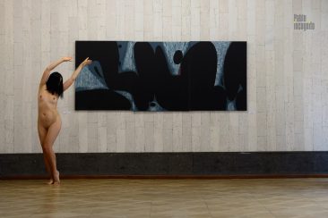 Naked with raised arms posing in front of a painting