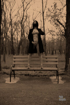 A woman in an unbuttoned raincoat stands on a bench, nude