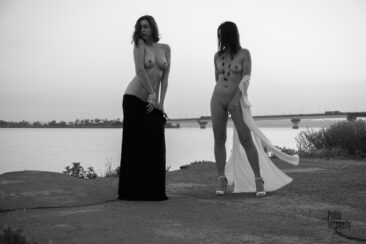 Two naked girls posing in front of a bridge