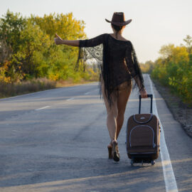 A girl in a cowboy hat and a cape on a naked body walks along the road with a suitcase