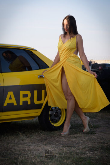 Iren Adler in a yellow dress near a retro Subaru nude