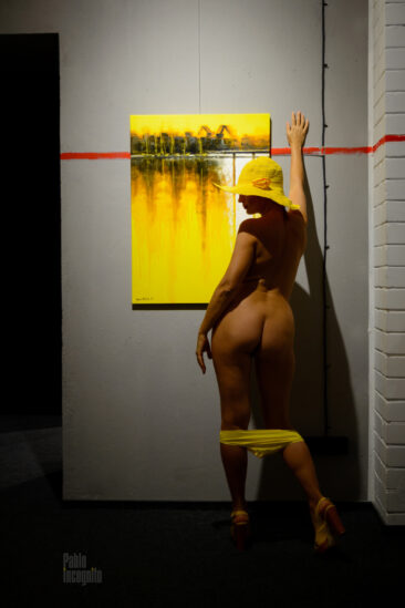 Naked in a yellow hat and shorts poses near a painting at an exhibition. Pablo Incognito