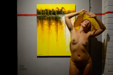Nude model poses naked near a yellow painting in a gallery. Photo by Pablo Incognito