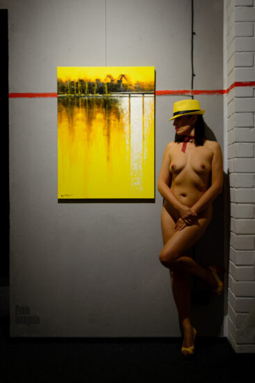 A nude model in a yellow hat and shoes poses naked near a painting in a gallery. Pablo Incognito